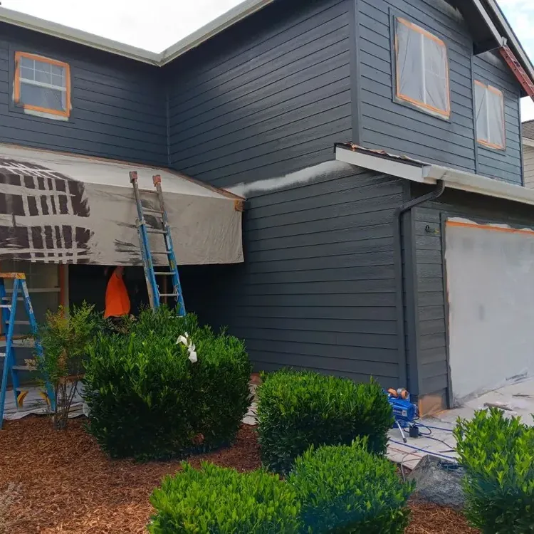 A two-story house mid-repaint in dark blue-grey, its windows and garage door masked, ladders against the wall