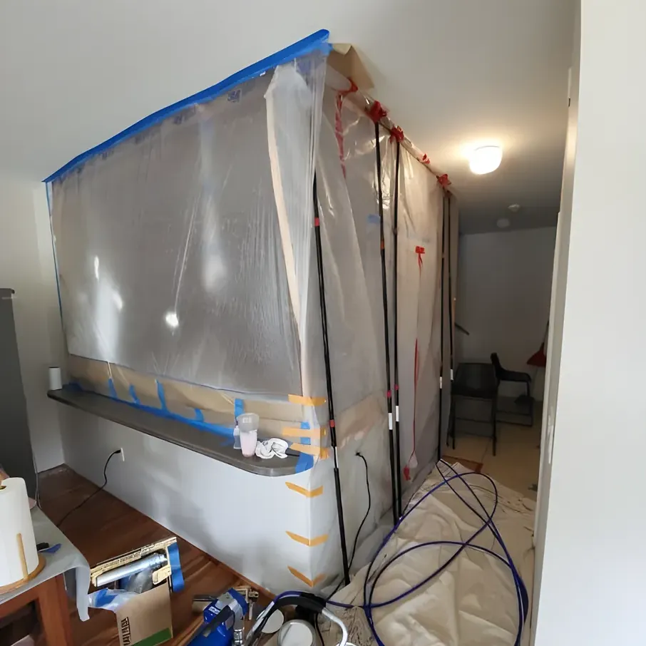 An interior wall sealed off in plastic sheeting and blue tape, with spray equipment on a covered floor