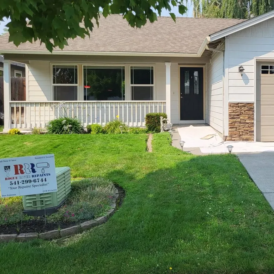 A cream single-story house with a white porch rail, repainted, with a Rogue River Repaints yard sign