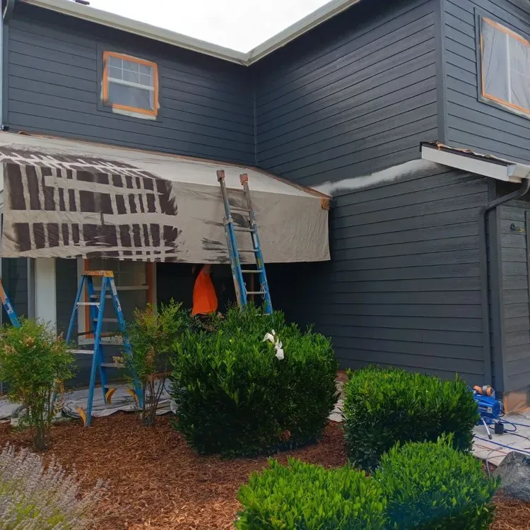 A two-story house mid-repaint in dark blue-grey, its windows and garage door masked
