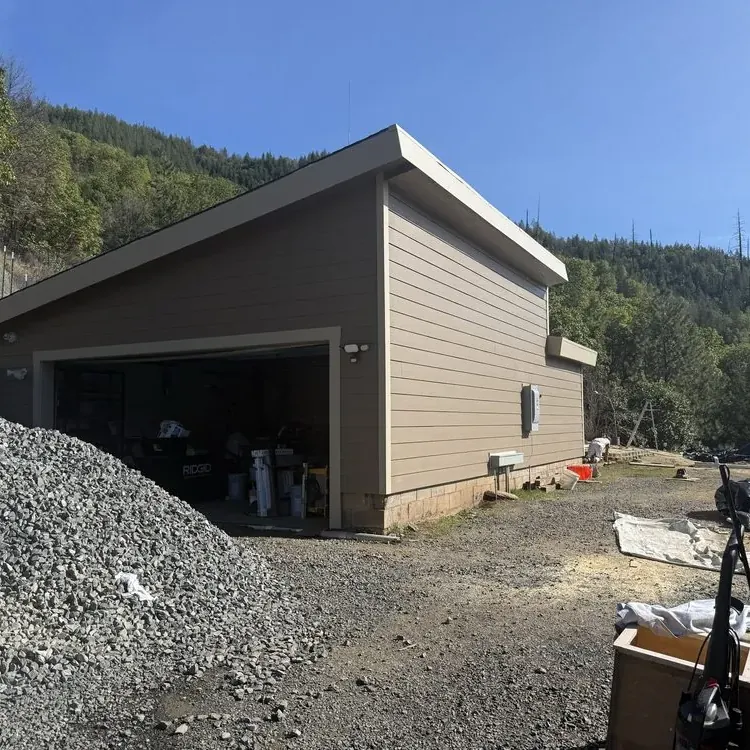 A detached garage re-sided in tan lap siding on a gravel lot below a wooded hillside