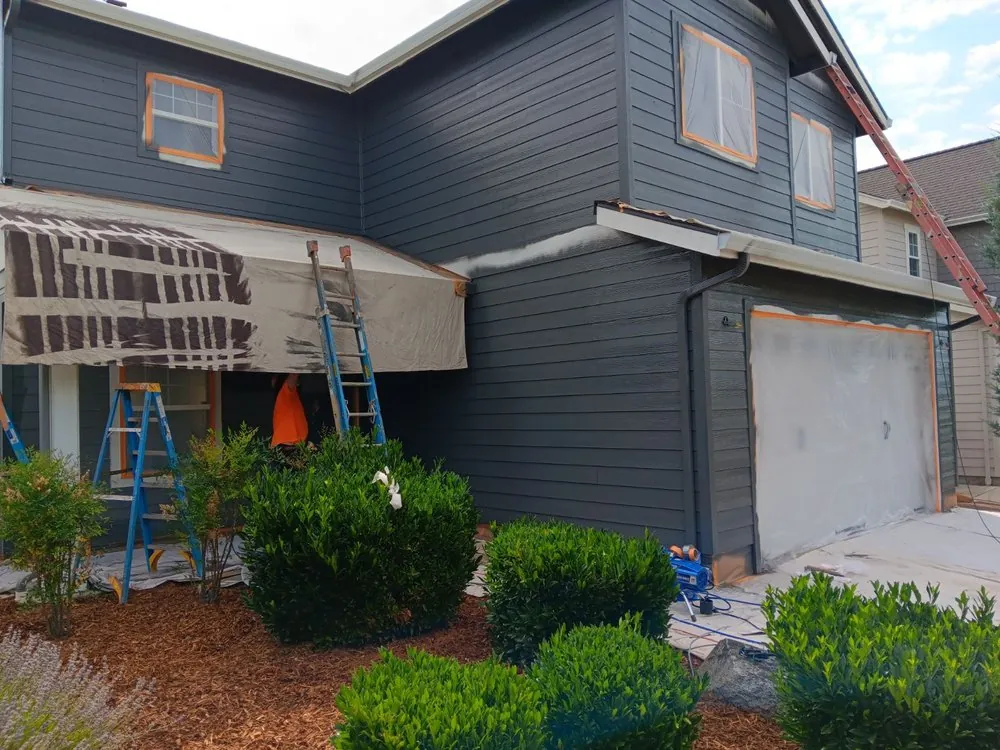 A two-story house mid-repaint in dark blue-grey, windows and garage door masked