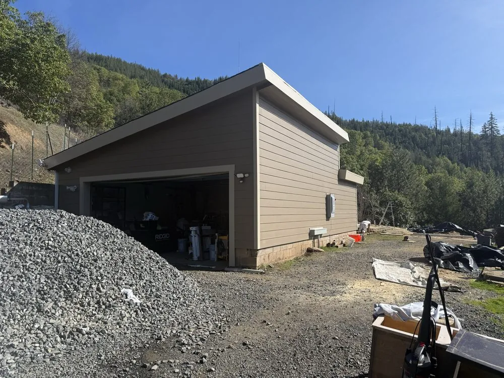A detached garage re-sided in tan lap siding on a gravel lot below a wooded hillside